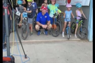 A group of children in colorful helmets and cycling gear are gathered with their bicycles, smiling for the camera. A man in a blue shirt is squatting in front of them. Banners in the background welcome participants to an event for mountain bike riders. There are cameras and equipment set up nearby, suggesting a photo or video shoot. Wee-chi-ta mountain bike trail.
