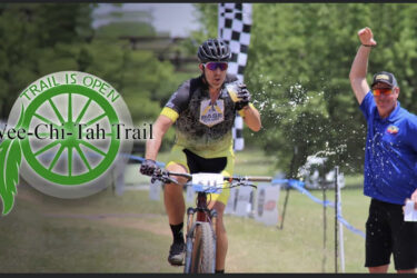 A cyclist in a yellow and black outfit splashes through water as he crosses the finish line, with arms raised in celebration. A man in a blue shirt smiles and cheers beside him. The image features a logo with the text "Trail is Open" and "Wee-Chi-Tah Trail." Behind them, a forested area and a checkered flag are visible, indicating the end of a race event. Wee-chi-ta mountain bike trail.