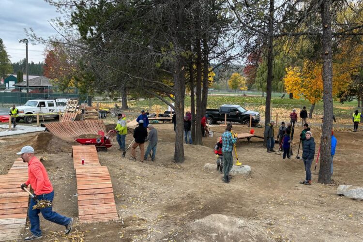 A group of volunteers working together at an outdoor park. They are building a wooden structure, likely a bike or skate ramp, amid trees with autumn foliage. Some individuals are using tools while others are transporting materials. The scene shows a collaborative effort in a natural setting, with vehicles and equipment in the background.