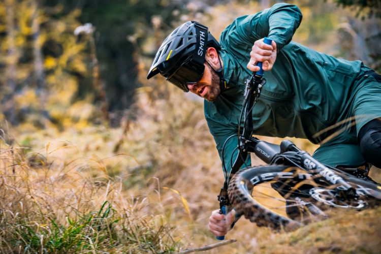 A mountain biker leans low while navigating a curve on a trail, surrounded by tall grass and autumn foliage. He wears a helmet and goggles for protection, and is focused on maintaining balance and control of his bike.