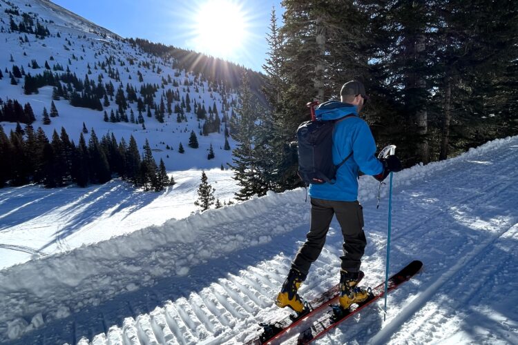 A skier ascending a snow-covered mountain trail, surrounded by trees, with the sun shining brightly in the background. The skier is wearing a blue jacket and yellow ski boots, carrying poles and a backpack. The landscape features a mix of snow, evergreen trees, and distant mountains.