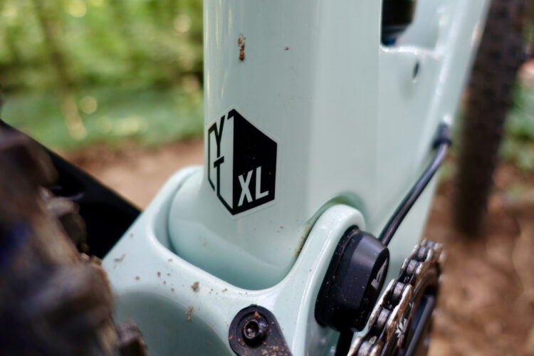 Close-up of a bicycle's frame featuring a light blue color and a visible logo that reads "YT XL." The image shows details of the bike's bottom bracket area, including dirt and debris, with parts of the chain and gear also partially visible. The background is blurred, indicating an outdoor setting.