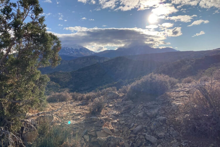 A scenic landscape featuring rocky terrain and sparse vegetation, with distant mountains partially covered by clouds. The sun is shining through the clouds, casting rays of light across the scene, highlighting the rugged natural beauty of the area.