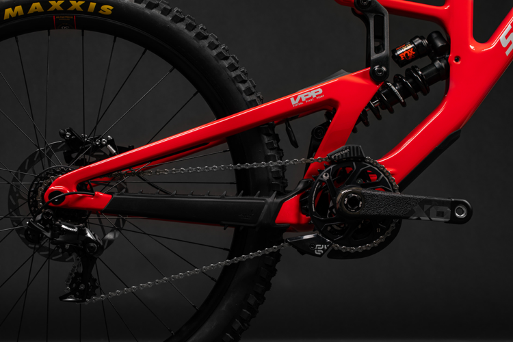 A close-up view of a red mountain bike frame showcasing the pedal, chain, and gearing components against a dark background. The image highlights the bike's VPP suspension system and features detailing on the crankset and rear derailleur.
