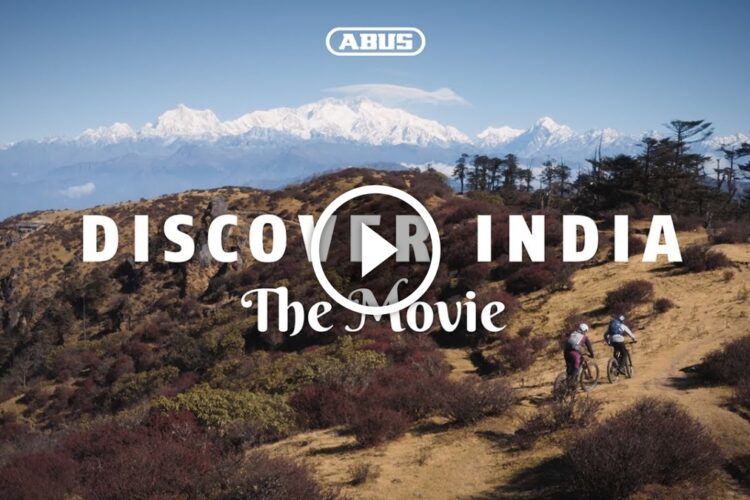 A picturesque mountain landscape in India with snow-capped peaks in the background. Two mountain bikers ride along a trail through a rugged terrain filled with colorful foliage. The image features the text "Discover India: The Movie" prominently displayed.