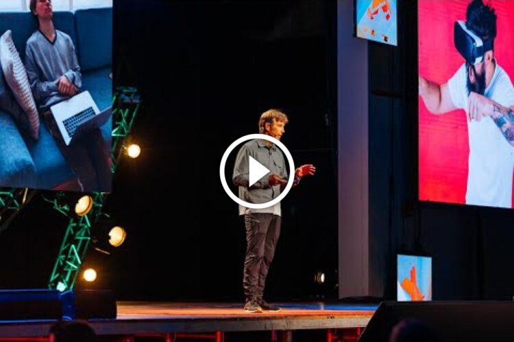 A speaker stands on stage presenting at an event. Two large screens display images: one of a person relaxing on a sofa with a laptop and another of a person wearing a virtual reality headset, engaged in an immersive experience. The stage is brightly lit with colorful lights.
