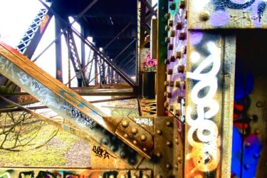 An industrial view of a partially graffiti-covered metal bridge structure. The image captures the intricate details of the bridge's supports and beams, showcasing colorful graffiti art on the rusted surfaces. Light filters through the gaps above, illuminating the scene. Springbank park mountain bike trail.