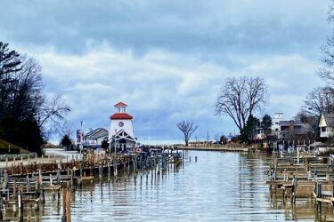 A tranquil waterfront scene featuring a calm canal lined with wooden docks and boat slips. In the distance, a white and red lighthouse-like structure stands near the water's edge, surrounded by bare trees and a cloudy sky. The scene reflects a peaceful atmosphere, evoking a sense of serenity in nature. The Beach mountain bike trail.