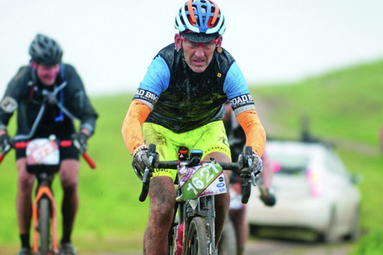 A muddy cyclist in bright gear competes in a mountain biking race, showing determination and focus. Another cyclist is visible in the background, and a vehicle can be seen parked nearby. The scene depicts challenging outdoor conditions.