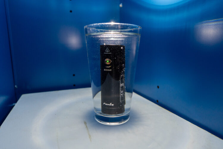 A glass filled with clear water, featuring a black flashlight submerged inside. The flashlight has a small green indicator light visible, and the background consists of a blue surface and wall.