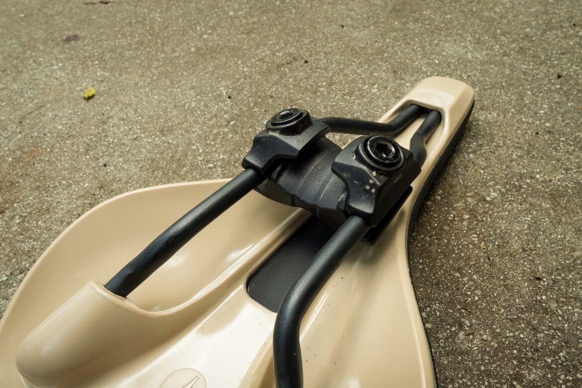 A close-up view of a beige SDG Bel Air mountain bike saddle, focusing on the black mounting hardware and support bars attached to the seat. The background shows a gray surface, possibly concrete, with a small leaf visible.