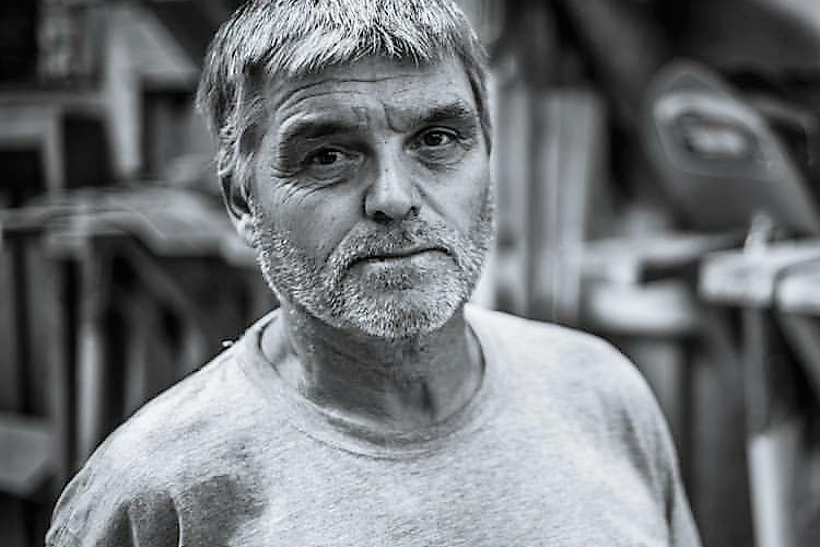 A man with short gray hair and a beard is looking directly at the camera, wearing a light gray t-shirt with the text "Gravity Logic" on it. The background is slightly blurred, depicting a workshop environment. The image is in black and white, emphasizing the man's thoughtful expression.