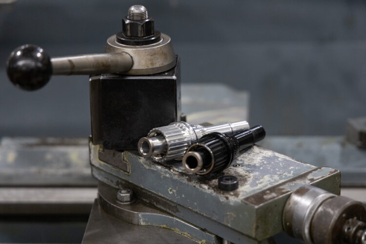 A close-up view of a machine component featuring a metallic control lever and two machine parts, one silver and one black, resting on a metallic surface. The background is blurred, highlighting the details of the machine parts and the textured surface of the machinery.