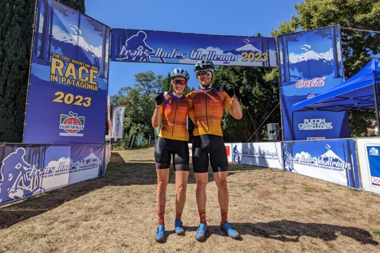 Alt text: Two athletes in cycling gear stand together at the finish line of the Trans Andes Challenge 2023 in Patagonia. They are smiling and giving a thumbs-up, with a colorful banner in the background showcasing the event.