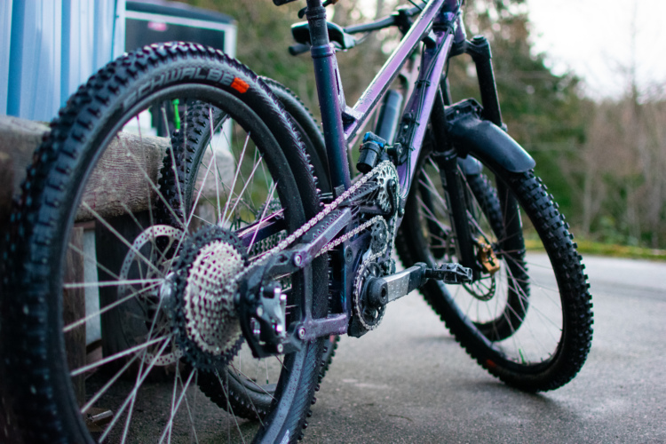 A close-up view of a mountain bike leaning against a wooden structure, showcasing its purple frame, detailed gears, and rugged tires. The pavement is visible beneath the bike, and the background features greenery, suggesting an outdoor setting.