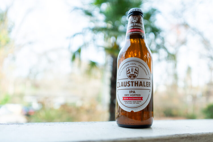 A close-up image of a Clausthaler Dry Hopped IPA non-alcoholic beer bottle sitting on a white surface, with a blurred outdoor background featuring greenery and soft natural light. The bottle showcases its label prominently, highlighting it as a non-alcoholic beverage with less than 0.5% alcohol by volume.