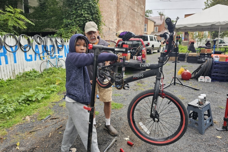 A young child with a focused expression stands next to an older man, both engaged in repairing a bicycle. The child is holding a bike repair stand, while the man assists in adjusting the bicycle. In the background, bicycles are leaning against a white fence with the words "GARDEN" visible. The scene takes place outdoors, under overcast skies, in a space filled with tools and bicycle parts.