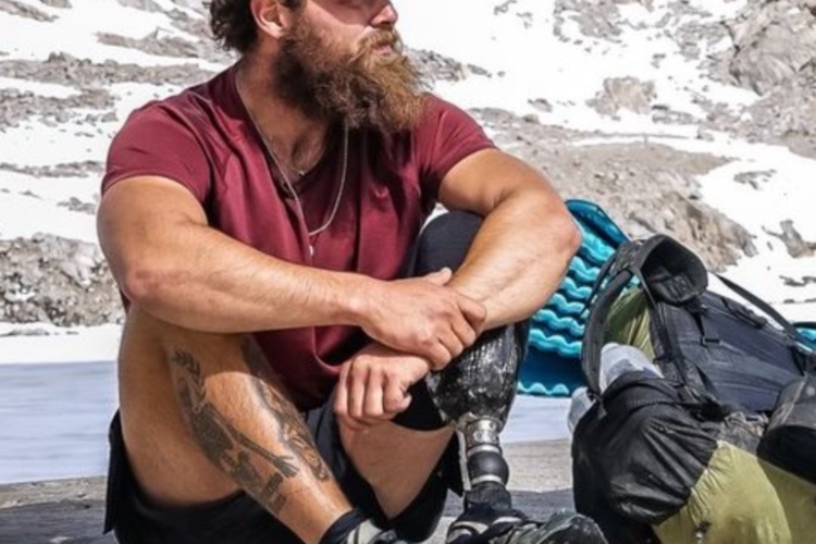 A man with a beard sits on a rocky surface near a snowy mountain backdrop. He is wearing a red shirt, shorts, and a cap, with a backpack beside him. His left leg features a prosthetic leg, and he has a detailed tattoo on his lower leg. The scene showcases a serene outdoor environment, emphasizing the man's contemplative expression.