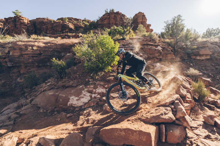 A mountain biker in black gear navigates a rocky trail, kicking up dust as they ride downhill. The background features red rock formations and sparse vegetation under a clear blue sky.