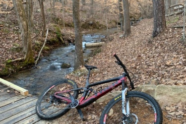 A mountain bike resting on the ground near a small creek in a forested area. The bike is positioned on a bed of fallen leaves, with trees in the background showing a mix of bare branches and scattered brown leaves. A wooden bridge is visible nearby, crossing over the creek. The scene has a serene, natural feel, typical of a walking or biking trail. Double Oak mountain bike trail.