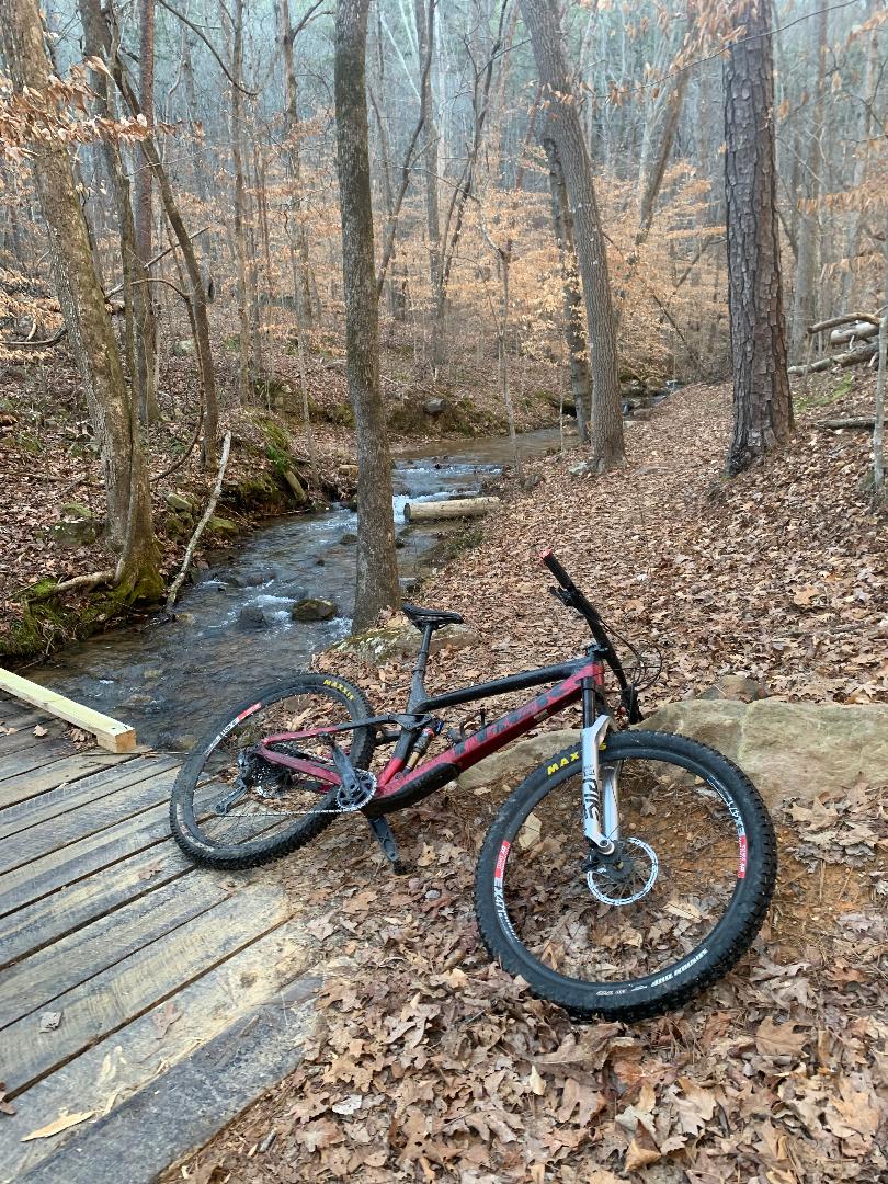 A mountain bike resting on the ground near a small creek in a forested area. The bike is positioned on a bed of fallen leaves, with trees in the background showing a mix of bare branches and scattered brown leaves. A wooden bridge is visible nearby, crossing over the creek. The scene has a serene, natural feel, typical of a walking or biking trail. Double Oak mountain bike trail.