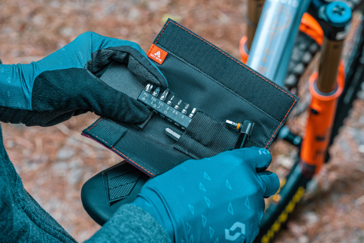A person wearing gloves holds an open tool pouch containing various bike repair tools, including a multi-bit screwdriver and several screwdriver bits. In the background, part of a mountain bike is visible, set against a natural outdoor setting.