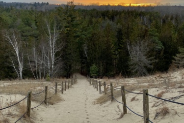 A sandy path leading down through a wooded area, bordered by wooden posts and rope. In the distance, the sun is setting, casting an orange glow against a cloudy sky, while green trees line the path on either side. Pinery Provincial Park mountain bike trail.
