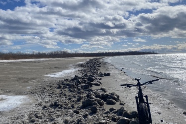 A black bicycle is parked on a sandy shore lined with rocky formations, with a backdrop of a cloudy blue sky. The water is visible on one side, and the landscape features patches of ice and a tranquil atmosphere. Presquile Provincial Park mountain bike trail.