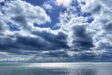 A serene beach scene featuring a sandy shore with scattered ice remnants. The calm water reflects the bright sun partially obscured by fluffy clouds, while rays of light break through the cloud cover, illuminating the sky in shades of blue and gray. Presquile Provincial Park mountain bike trail.