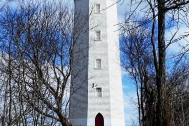A tall white lighthouse with a red top is partially surrounded by bare trees, set against a bright blue sky with fluffy clouds. Snow covers the ground in the foreground, leading up to the lighthouse, which features a red door and several small windows. The scene conveys a serene winter atmosphere by the water. Presquile Provincial Park mountain bike trail.