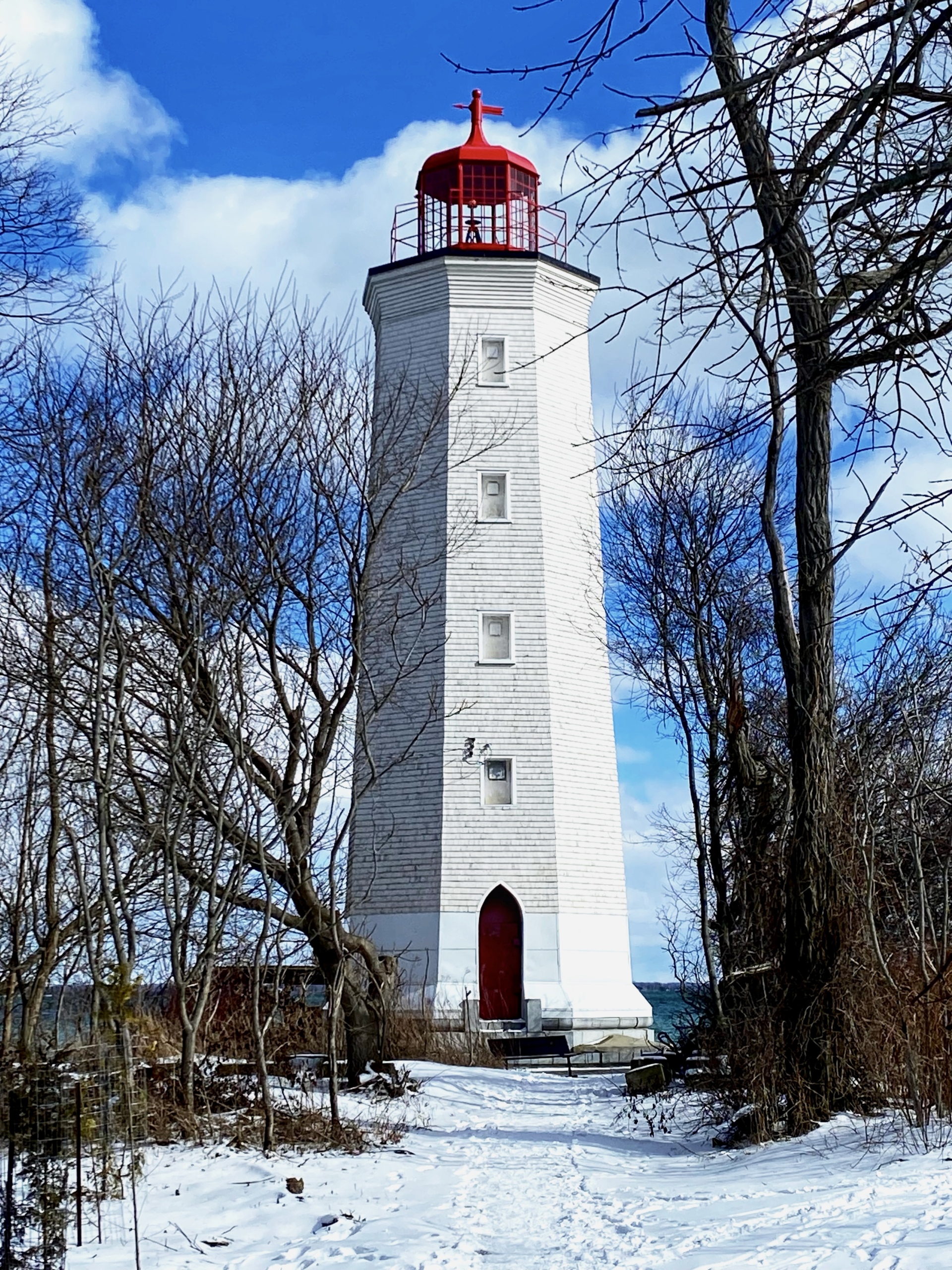 A tall white lighthouse with a red top is partially surrounded by bare trees, set against a bright blue sky with fluffy clouds. Snow covers the ground in the foreground, leading up to the lighthouse, which features a red door and several small windows. The scene conveys a serene winter atmosphere by the water. Presquile Provincial Park mountain bike trail.