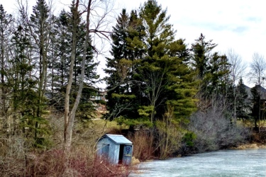 A serene winter scene featuring a partially frozen lake surrounded by trees. A small, weathered shed is nestled among shrubs along the water's edge, under a cloudy sky. MeadowLilly mountain bike trail.