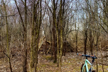 A mountain bike stands on a dirt path surrounded by sparse trees and underbrush in a wooded area. The scene is captured in late winter or early spring, with some fallen leaves on the ground and bare branches overhead. The sky is overcast, hinting at a cool day. MeadowLilly mountain bike trail.