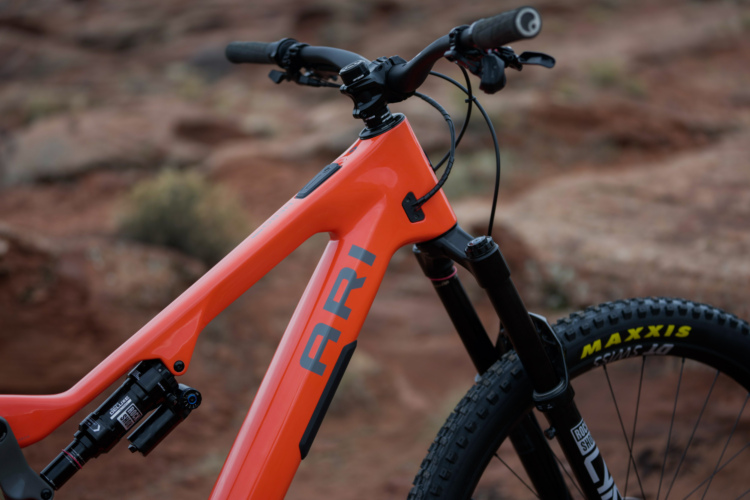 Close-up of an orange Ari mountain bike frame, featuring the word "ARI" visibly displayed along the side. The image captures the handlebars, front fork, and part of the wheel, set against a background of rocky terrain.