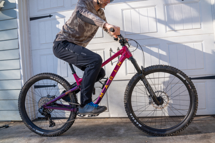 A person wearing a camouflage long-sleeve shirt and dark pants is riding a purple mountain bike, positioned beside a garage door. The bike features thick tires and a sleek frame, highlighting its suitability for off-road terrain.