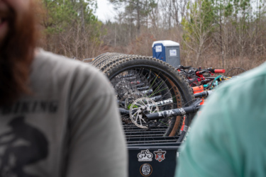 Two individuals are partially visible in the foreground, with a focus on their shoulders. In the background, a collection of mountain bikes is stacked on a vehicle. The image also includes a portable restroom and trees in the surrounding area, indicating an outdoor setting. Jarrod's Place mountain bike trail.