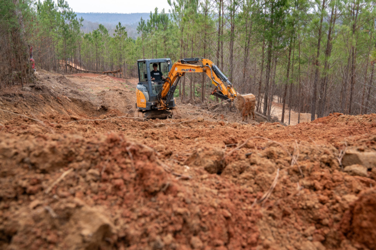 A yellow excavator working on a dirt path in a wooded area, with piles of freshly dug soil in the foreground and trees in the background.