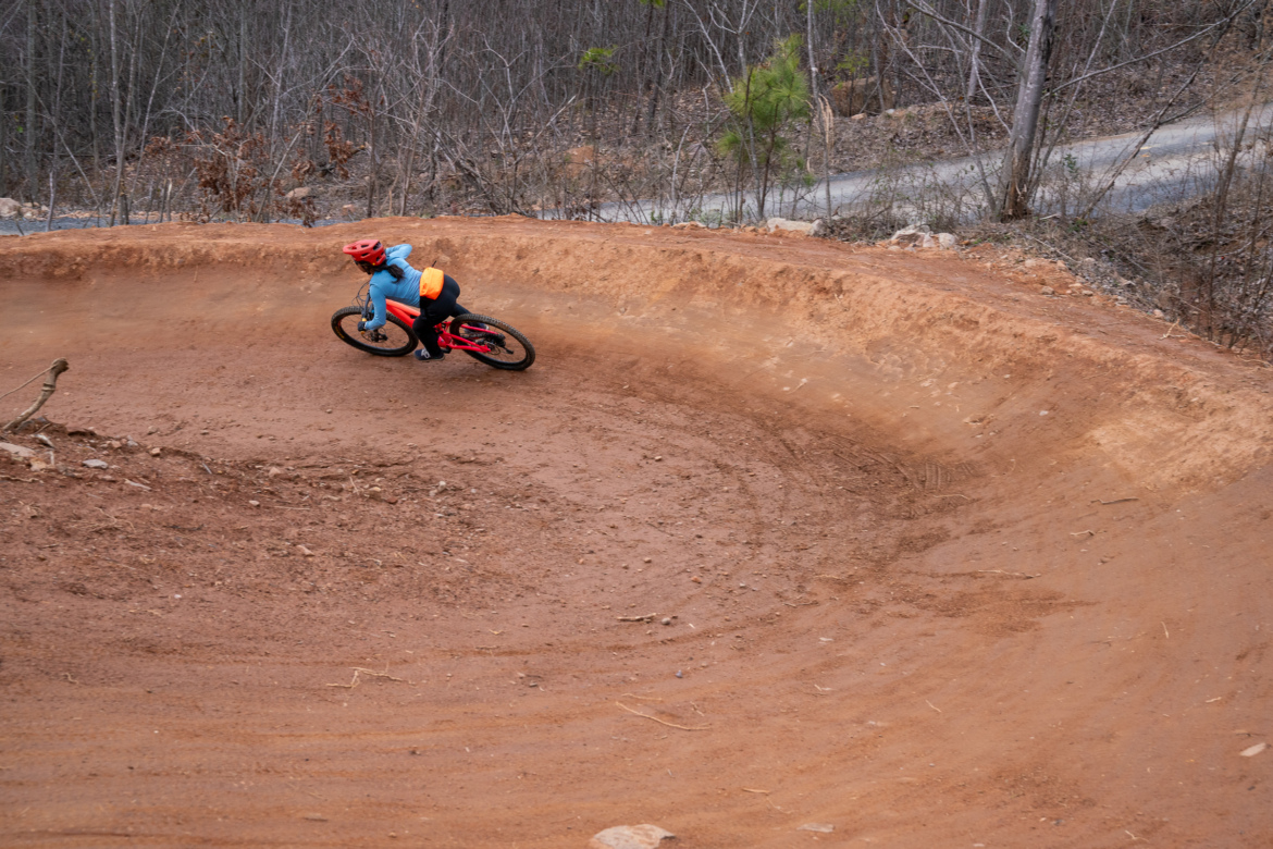 A cyclist in a blue jacket and red helmet leans into a tight turn on a dirt bike trail surrounded by trees in a natural setting. The trail is sandy, and a winding path is visible in the background.