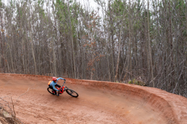 A mountain biker wearing a red helmet and blue long-sleeve shirt navigates a dirt trail, leaning into a curve surrounded by tall trees. Jarrod's Place mountain bike trail.