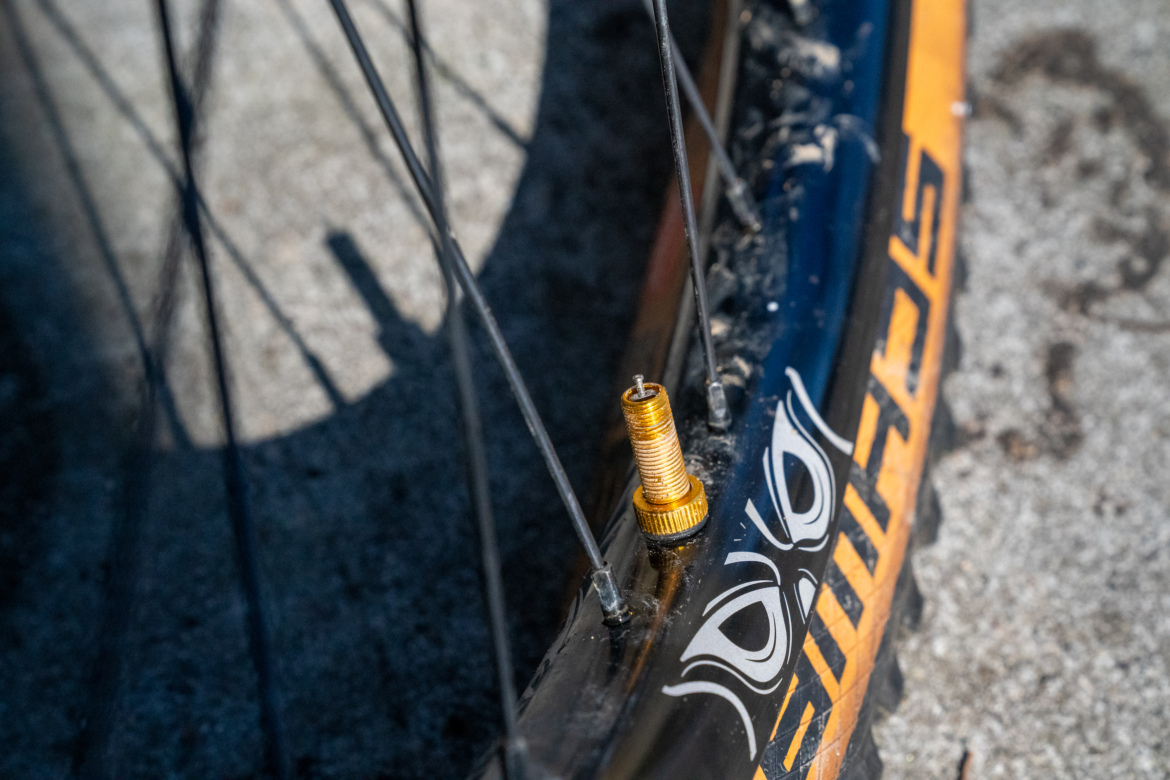 Close-up image of a bike tire showing the valve stem, which is metallic gold in color, with spokes visible in the background. The tire has a detailed design in black and orange, and the ground surface appears textured and gray.
