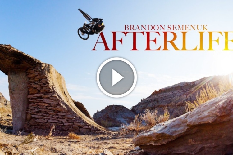 A mountain biker performing a mid-air stunt over a rocky ledge in a desert landscape, with the title "Brandon Semenuk AFTERLIFE" prominently displayed above the action. The sun shines brightly in the background, enhancing the outdoor ambiance. A play button icon indicates that this image is part of a video.