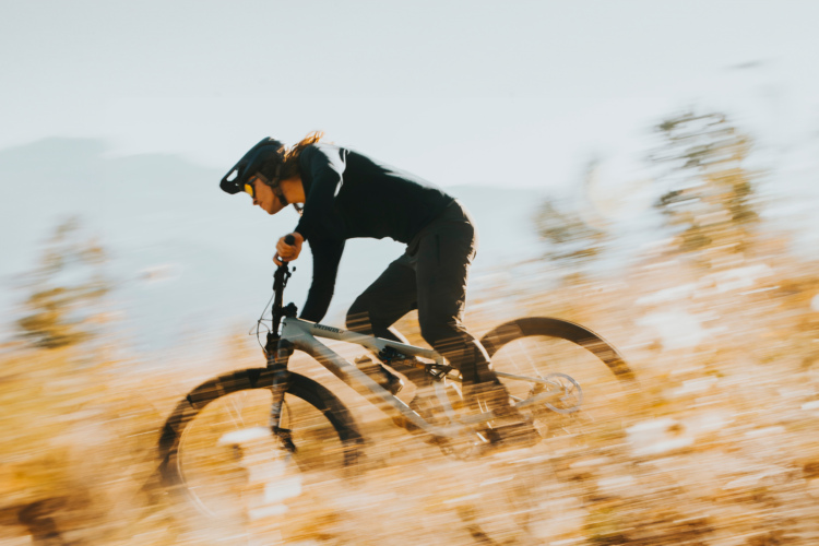 A person riding a mountain bike at high speed through a field of tall grass, with blurred motion effects illustrating movement. The rider is wearing a helmet and dark athletic clothing, showcasing an energetic outdoor landscape.