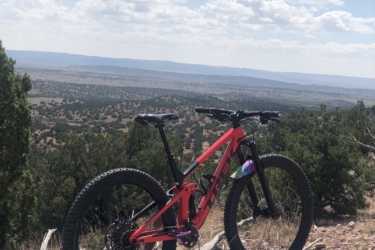A vibrant pink mountain bike rests against a wooden stump on a dirt trail, surrounded by sparse greenery and a vast landscape under a blue sky with scattered clouds. The view extends across rolling hills and valleys in the distance, showcasing the beauty of nature. Galisteo Basin Preserve mountain bike trail.