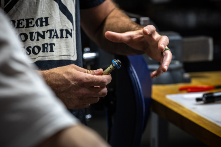 A close-up of hands demonstrating a detailed discussion or analysis of a small mechanical component, likely in a workshop environment. One hand holds a cylindrical piece with visible markings, while the other gestures expressively, indicating explanation or instruction. Tools and materials are subtly visible in the background, creating a focused, hands-on atmosphere.