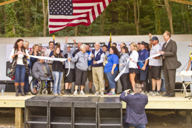 A large group of people gathers on a wooden stage for a ribbon-cutting ceremony. They are celebrating with smiles and raised hands, while an American flag hangs overhead. Among the crowd, a man stands in the center holding a pair of scissors, ready to cut the ribbon. There are individuals in wheelchairs and various attire, reflecting a diverse community. A photographer captures the moment from the front. Griffin Bike Park mountain bike trail.