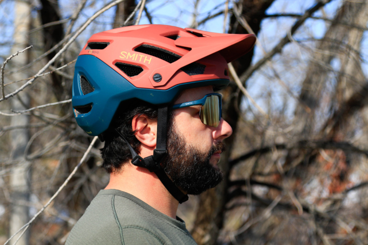 A man with a beard wearing a colorful bike helmet and sunglasses, facing sideways against a backdrop of bare branches. The helmet features a combination of coral and blue colors, with the brand name visible.