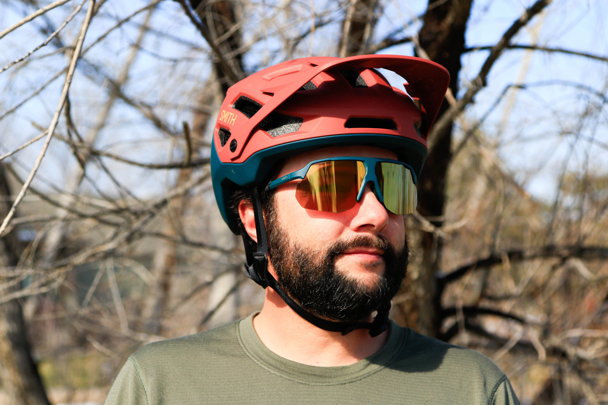 A person wearing a red and teal mountain biking helmet and reflective sunglasses stands outdoors, with a backdrop of bare trees. The individual has a beard and is wearing a green t-shirt.
