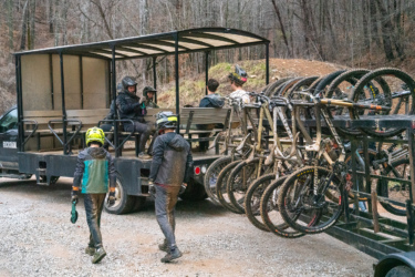 A group of mountain bikers, wearing helmets and muddy gear, are gathered around an open trailer equipped to transport bikes. In the trailer, several dirty mountain bikes are displayed, with some riders sitting down while others walk nearby on a gravel path surrounded by trees. Windrock Bike Park mountain bike trail.