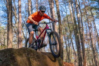 A person in an orange shirt and black shorts rides a mountain bike over a large rock in a wooded area. The scene features tall trees and blue skies, highlighting an outdoor adventure in nature. Lakeshore Bike Park mountain bike trail.