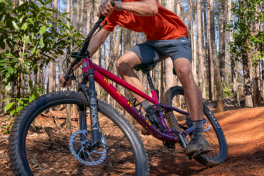 A person riding a mountain bike on a dirt trail in a wooded area, wearing a helmet and sunglasses. The rider is leaning into a turn, showcasing an action shot with trees and foliage in the background. Lakeshore Bike Park mountain bike trail.